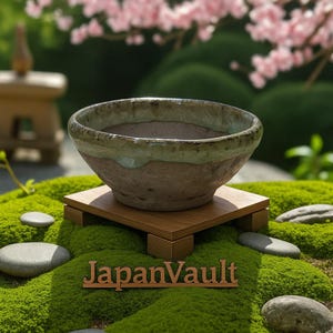 以下が含まれることがあります： 緑と茶色の釉薬を施した陶器のボウルが、小さな木のプラットフォームに置かれています。ボウルは、緑の苔、石、そしてピンクの花が咲くぼやけた庭のシーンを背景に展示されています。「JapanVault」の文字が見えます。