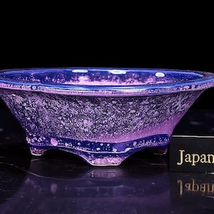 Puede incluir: Una maceta de bonsái ovalada hecha a mano con un esmalte moteado púrpura y azul. La maceta de cerámica tiene tres pequeños pies y un borde azul oscuro. El texto "Japan Vault" está en una etiqueta negra.