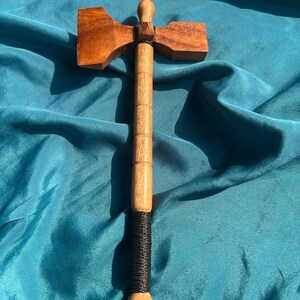 May include: A wooden tomahawk featuring a polished brown axe head and a long, cylindrical handle. The handle is segmented with horizontal grooves and includes a black cord wrap at the base, with additional black cord securing the axe head to the shaft.