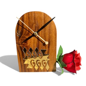 May include: Wooden desk clock with gold-colored hands and a decorative gold-colored design. The clock is next to a red rose. The clock's face has the number 9. The clock is made of wood.