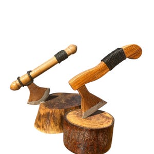 May include: Two miniature wooden axes with brown handles and silver-colored blades. The handles are wrapped with black twine. The axes are displayed on small, round wooden blocks, showcasing a rustic aesthetic.