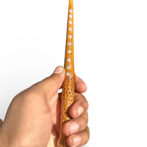 May include: A light brown wooden wand with a textured handle and a tapered tip. The wand features small, clear crystals embedded along its length. The wand is held in a hand against a white background.