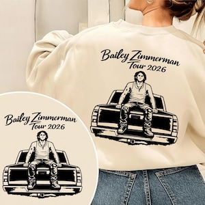 Peut inclure: Sweat-shirt crème avec un graphisme noir d'une personne assise sur une benne de camion, et le texte "Bailey Zimmerman Tour 2026" en écriture cursive. Le motif est répété sur un petit graphisme circulaire.