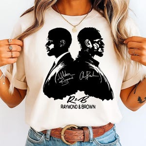May include: A cream-colored t-shirt featuring a black silhouette design of two men facing each other. The design includes signatures and the text "R&B RAYMOND & BROWN" in a bold font. The shirt is made of a soft material.