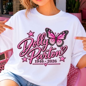 May include: A white t-shirt featuring a graphic design with the text Dolly Parton 1946-2026 in pink glittery script. The design includes a pink and black butterfly, several pink stars, and small pink hearts surrounding the text elements.