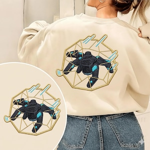 Könnte beinhalten: Cremefarbenes Sweatshirt mit einem gestickten Design einer schwarz-blauen Roboter-Spinne in einem goldenen geometrischen Rahmen. Das Design befindet sich auf der Rückseite und als kleiner Aufnäher.