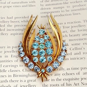 Könnte beinhalten: Vintage goldfarbene Brosche mit floralem Design. Die Brosche hat einen zentralen Cluster aus hellblauen Strasssteinen, umgeben von einem Ring aus ähnlichen Steinen und goldfarbenen Blättern. Unten befindet sich ein Sternmuster.