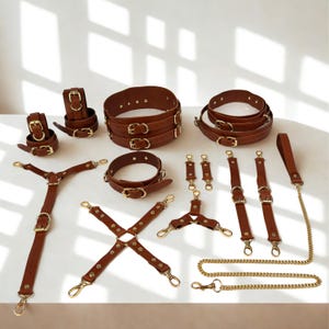 Pode incluir: Um conjunto de acessórios em couro castanho com detalhes dourados. O conjunto inclui cintos, algemas, suspensórios e uma trela com corrente. Os itens estão dispostos em uma superfície branca, mostrando os vários designs.