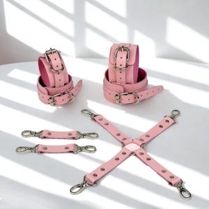 Puede incluir: Juego de sujeciones de cuero rosa, que incluye esposas, tobilleras y correas de conexión. El juego presenta hebillas y herrajes de metal plateado. Los puños están forrados con un material rosa más oscuro.