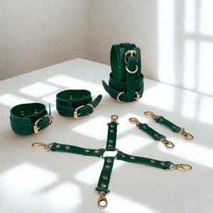 May include: A collection of green leather restraints with gold-colored hardware. The set includes wrist cuffs, an ankle cuff, and a multi-strap harness. The items are arranged on a white surface, showcasing the details of the buckles and clasps.