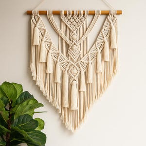 May include: Cream-coloured macrame wall hanging with a geometric design. The piece features intricate knotting, diamond patterns, and multiple tassels. It is suspended from a natural wooden dowel, adding a touch of bohemian style to any space.