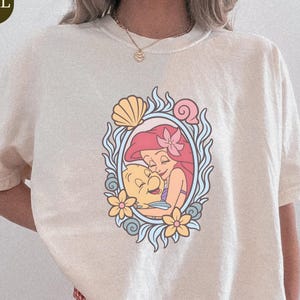 A person wearing a white t-shirt with a graphic of a mermaid and a flower design.