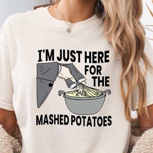 Puede incluir: Camiseta color crema con la frase "I'M JUST HERE FOR THE MASHED POTATOES". El diseño muestra una mano con traje removiendo puré de patatas. Parte de una colección de ropa.