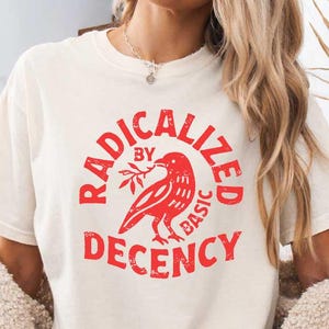 以下が含まれることがあります： 「RADICALIZED BY BASIC DECENCY」と書かれた赤いグラフィックが特徴のクリーム色のTシャツ。 グラフィックは枝を持った鳥を描いています。 このTシャツはアパレルコレクションの一部です。