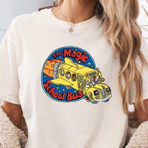 May include: A cream-coloured t-shirt with a colourful graphic of the Magic School Bus. The design shows a yellow school bus with a rocket engine, the text "The Magic School Bus" in a blue circle, and the word "NATURAL" in a brown oval.