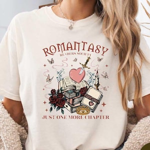 Könnte beinhalten: Cremefarbenes T-Shirt mit dem Text "ROMANTASY READERS SOCIETY" und einer Grafik aus Büchern, einem Herz, einem Schwert und Schmetterlingen. Der Satz "JUST ONE MORE CHAPTER" steht darunter. Das Shirt hat einen Rundhalsausschnitt.