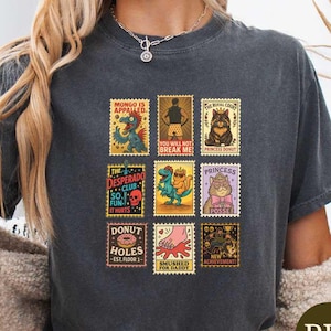May include: Dark gray t-shirt featuring nine colorful postage stamp designs with various illustrations and text. The stamps include images of a dinosaur, cat, and other whimsical characters. The shirt is labeled "Pepper" at the bottom.