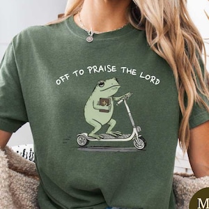 Può includere: T-shirt verde militare con la scritta "OFF TO PRAISE THE LORD". La maglietta presenta un cartone animato di una rana che va in scooter e tiene in mano un libro. La maglietta è di uno stile comodo e casual.