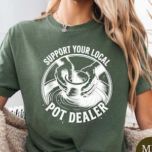 May include: Military green t-shirt with a white graphic that reads "Support Your Local Pot Dealer" in a circular design. The design features hands shaping a pot on a pottery wheel. The shirt is a comfortable, casual style.