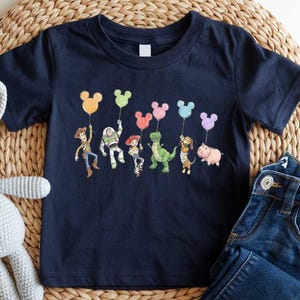 A navy blue t-shirt with a graphic of cartoon characters and balloons, a gray crocheted bunny, and a pair of blue jeans are arranged on a woven placemat.