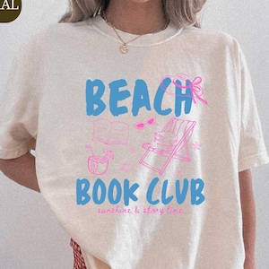 May include: Cream-colored t-shirt with the words "BEACH BOOK CLUB" in blue and pink, along with beach-themed illustrations. The shirt also has the words "sunshine & story time" in a smaller font. The word "NATURAL" is in a green box.