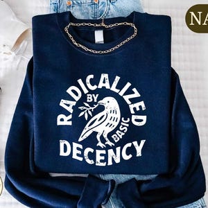 May include: Navy blue sweatshirt with the text "RADICALIZED BY BASIC DECENCY" in a circular design. A white dove holding a sprig is in the center. A gold chain necklace is draped on the sweatshirt. White sneakers are visible.