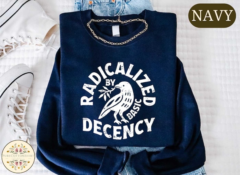 May include: Navy blue sweatshirt with the text "RADICALIZED BY BASIC DECENCY" in a circular design. A white dove holding a sprig is in the center. A gold chain necklace is draped on the sweatshirt. White sneakers are visible.