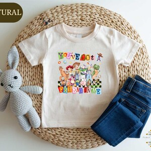 A white t-shirt with a colorful graphic design, a crocheted stuffed rabbit, and a pair of blue jeans are arranged on a woven mat.