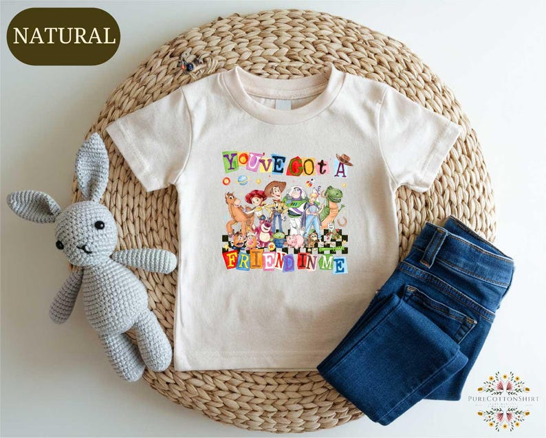 A white t-shirt with a colorful graphic design, a crocheted stuffed rabbit, and a pair of blue jeans are arranged on a woven mat.