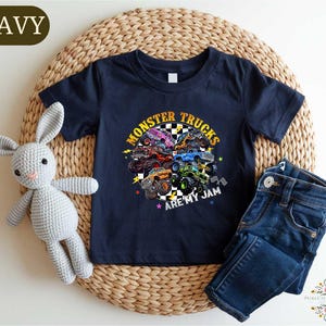 A navy blue t-shirt with a graphic of monster trucks and the text "MONSTER TRUCKS ARE MY JAM" printed on it, along with a pair of blue jeans and a crocheted stuffed rabbit.