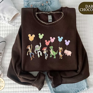 a brown sweatshirt with a cartoon character design, including a group of characters such as Woody, Buzz Lightyear, and Piggy, along with various other characters from the Toy Story franchise. The sweatshirt is laid out on a bed, and there are a pair of sneakers and a pair of jeans visible in the background.