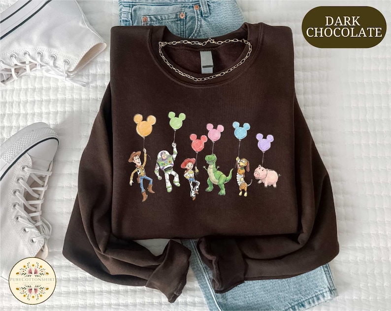 a brown sweatshirt with a cartoon character design, including a group of characters such as Woody, Buzz Lightyear, and Piggy, along with various other characters from the Toy Story franchise. The sweatshirt is laid out on a bed, and there are a pair of sneakers and a pair of jeans visible in the background.