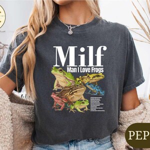 A woman wearing a gray t-shirt with a graphic design of various colored frogs.
