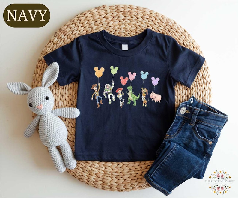A navy blue t-shirt with a graphic of cartoon characters and balloons, a gray crocheted bunny, and a pair of blue jeans are arranged on a woven placemat.