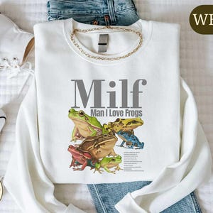 a white t-shirt with a graphic design of a frog and the text "Milf Man I Love Frogs" printed on it, along with a pair of jeans and white sneakers.