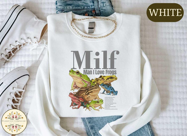 a white t-shirt with a graphic design of a frog and the text "Milf Man I Love Frogs" printed on it, along with a pair of jeans and white sneakers.