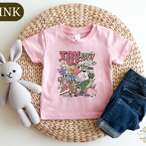 A pink t-shirt with a cartoon character design, a crocheted stuffed rabbit, and a pair of blue jeans are arranged on a woven placemat.