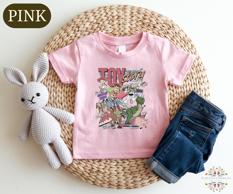 A pink t-shirt with a cartoon character design, a crocheted stuffed rabbit, and a pair of blue jeans are arranged on a woven placemat.