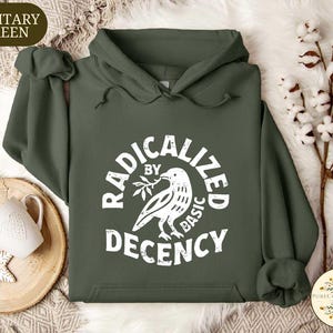 May include: Military green hoodie with a white graphic that reads "RADICALIZED BY BASIC DECENCY" around a dove holding an olive branch. The hoodie has a drawstring hood and a front pocket. A white mug and snowflake ornament are on a wooden tray.