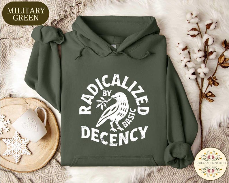 May include: Military green hoodie with a white graphic that reads "RADICALIZED BY BASIC DECENCY" around a dove holding an olive branch. The hoodie has a drawstring hood and a front pocket. A white mug and snowflake ornament are on a wooden tray.