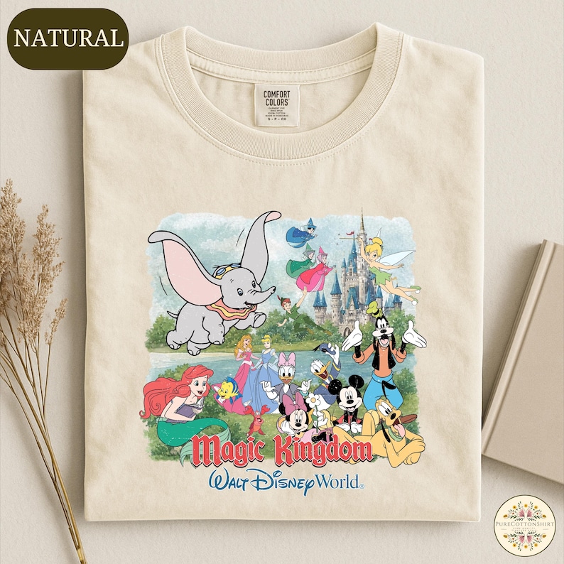 A t-shirt with a colorful graphic design depicting various Disney characters from the Magic Kingdom, including Dumbo, Minnie Mouse, Mickey Mouse, Donald Duck, and Goofy, along with a castle and a lake.