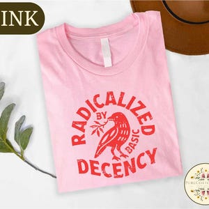 May include: A light pink t-shirt with the words "RADICALIZED BY BASIC DECENCY" in red, along with a red bird graphic. The shirt has a crew neck and a white tag. A brown hat is visible in the background.