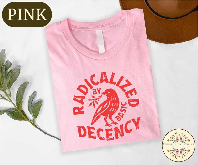 May include: A light pink t-shirt with the words "RADICALIZED BY BASIC DECENCY" in red, along with a red bird graphic. The shirt has a crew neck and a white tag. A brown hat is visible in the background.
