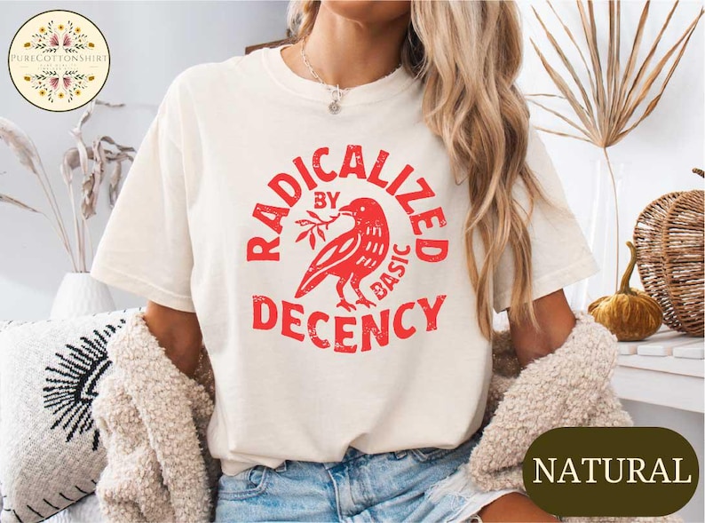 May include: Cream-colored t-shirt with a red graphic that reads "RADICALIZED BY BASIC DECENCY." The graphic features a bird holding a branch. The shirt is part of a clothing collection.