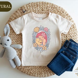 A white t-shirt with a mermaid design, a crocheted gray rabbit, and a pair of blue jeans are arranged on a woven placemat.