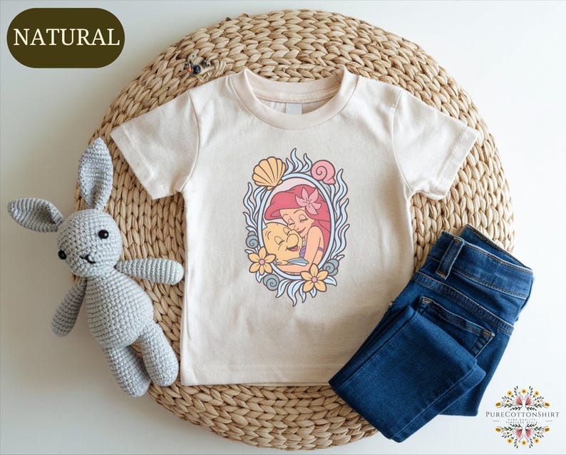 A white t-shirt with a mermaid design, a crocheted gray rabbit, and a pair of blue jeans are arranged on a woven placemat.
