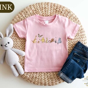 A pink t-shirt with a line of cartoon animal characters printed on it, a crocheted gray bunny, and a pair of blue jeans are arranged on a woven placemat.