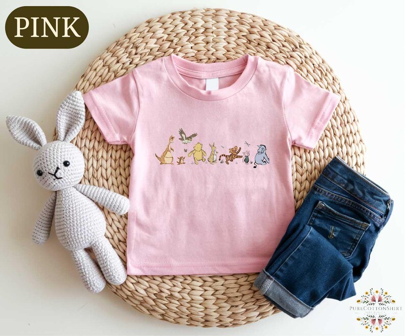 A pink t-shirt with a line of cartoon animal characters printed on it, a crocheted gray bunny, and a pair of blue jeans are arranged on a woven placemat.