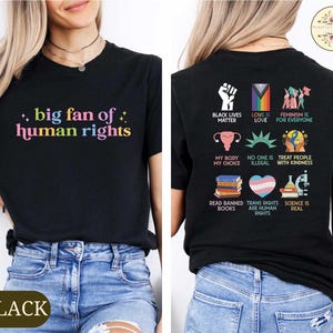 May include: Black t-shirt with the text "big fan of human rights" in colorful letters. The back of the shirt features various social justice slogans and symbols, including "Black Lives Matter" and "Trans Rights Are Human Rights".