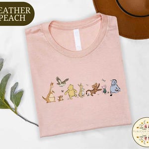A pink t-shirt with a line of colorful cartoon animal characters printed on it, including a kangaroo, lion, tiger, and elephant, along with a brown hat and some greenery.